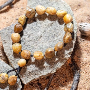 May include: A beaded bracelet made of rounded, amber-colored stones. The bracelet is strung on a black cord and rests on a gray stone, with a sandy background. The stones vary in size and shade, creating a natural, earthy aesthetic.