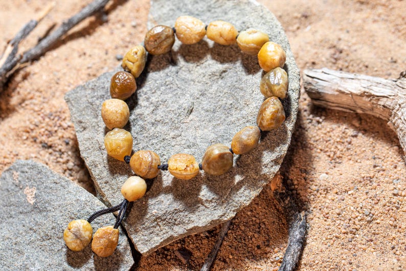 May include: A beaded bracelet made of rounded, amber-colored stones. The bracelet is strung on a black cord and rests on a gray stone, with a sandy background. The stones vary in size and shade, creating a natural, earthy aesthetic.