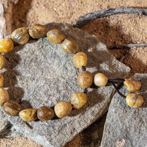 May include: A beaded bracelet made of light brown, irregularly shaped beads. The beads are strung on a black cord and rest on a gray stone. The background is a sandy texture.