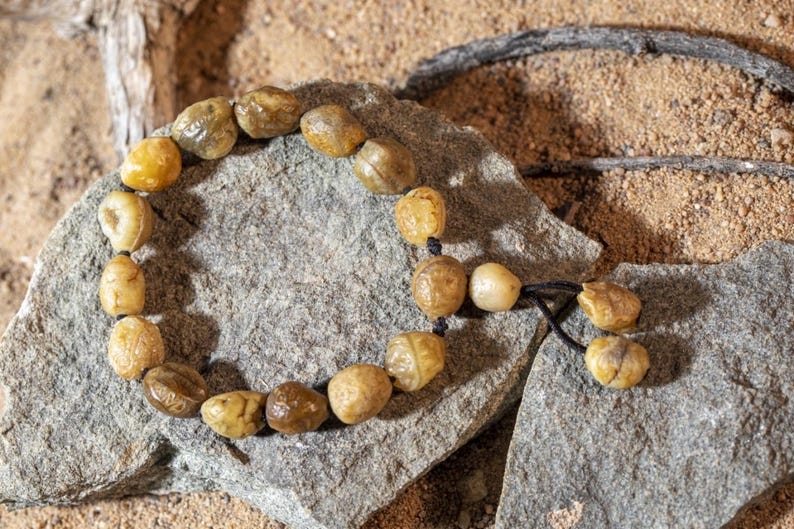 May include: A beaded bracelet made of light brown, irregularly shaped beads. The beads are strung on a black cord and rest on a gray stone. The background is a sandy texture.