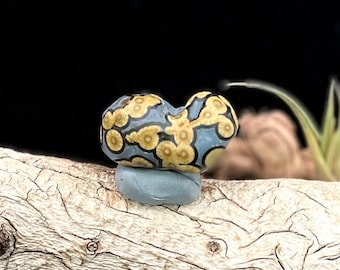 May include: A unique, handcrafted bead with a blue base and a pattern of yellow and black circles. The bead is resting on a small, light blue stone, and is displayed on a piece of driftwood.
