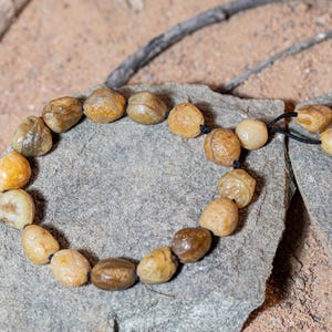 May include: A beaded bracelet made of smooth, rounded stones in shades of tan, brown, and cream. The stones are strung on a black cord and rest on a gray stone surface. The bracelet is a natural, earthy accessory.