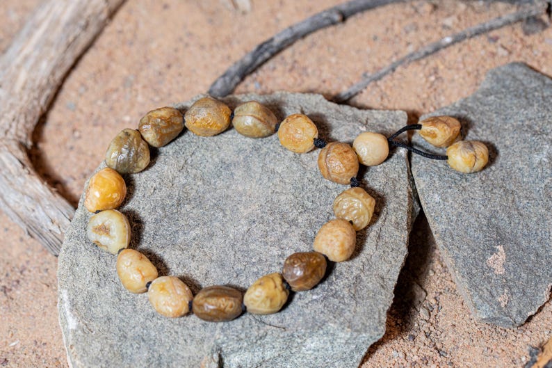 May include: A beaded bracelet made of smooth, rounded stones in shades of tan, brown, and cream. The stones are strung on a black cord and rest on a gray stone surface. The bracelet is a natural, earthy accessory.