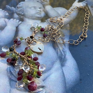 May include: A gold chain necklace with a heart-shaped locket and a dangling charm featuring a variety of gemstones, including clear, green, and red.