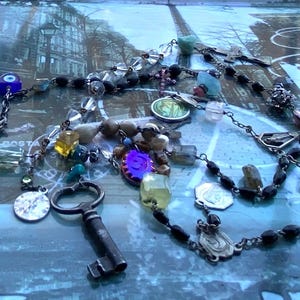 May include: A silver chain necklace with a variety of charms, including a key, a small religious medal, and a blue evil eye bead. The necklace is on a blue and white patterned surface.