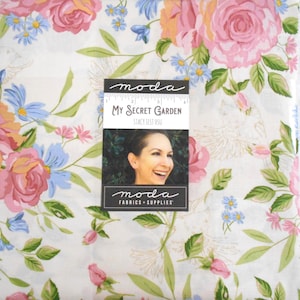 May include: A white fabric featuring a floral pattern of pink roses, blue flowers, and green leaves. A rectangular label is centered on the fabric, displaying the text "moda", "My Secret Garden", "Stacy Iest Hsu", and a 4-inch ruler scale.