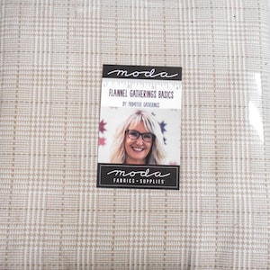 May include: A folded piece of light beige and white plaid flannel fabric. A rectangular product label is attached to the center, featuring the text "moda FLANNEL GATHERINGS BASICS BY: PRIMITIVE GATHERINGS" and a photograph of a person with glasses.