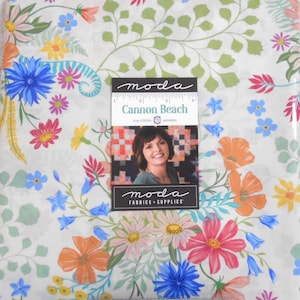 May include: A fabric panel with a floral design in shades of blue, pink, orange, and yellow. The fabric features a variety of flowers and green leaves on a white background. A label reads "Cannon Beach" by Crystal Manning.
