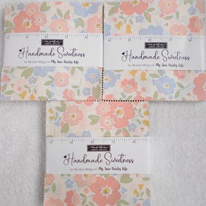 Puede incluir: Tres pilas de piezas de tela cuadradas con un estampado floral en tonos melocotón, azul claro y verde. Cada pila está envuelta en una banda de papel blanco con la etiqueta Handmade Sweetness de Heather Briggs de My Sew Quilty Life con una regla.