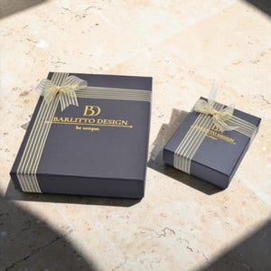 May include: Two dark gray gift boxes with gold ribbon and accents. The larger box has the text "BARLITTO DESIGN. be unique." printed in gold. The boxes are on a light-colored stone surface, with shadows.