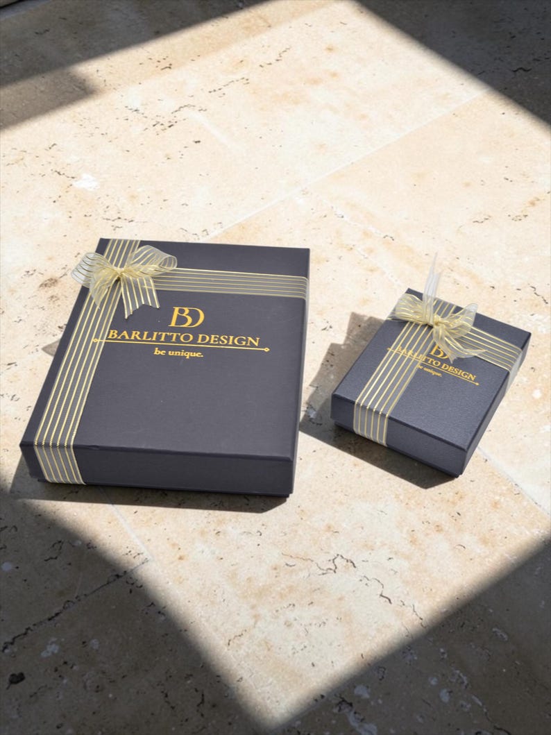 May include: Two dark gray gift boxes with gold ribbon and accents. The larger box has the text "BARLITTO DESIGN. be unique." printed in gold. The boxes are on a light-colored stone surface, with shadows.
