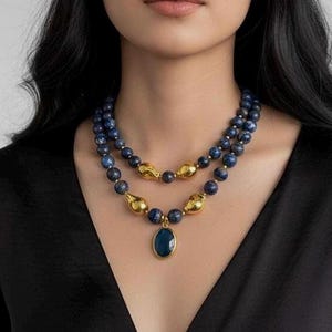 May include: A layered necklace featuring dark blue beads and gold-colored accents. The necklace has a pendant with a dark blue oval stone. The model is wearing a black top.