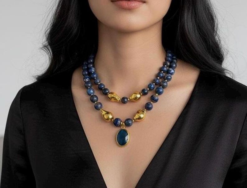 May include: A layered necklace featuring dark blue beads and gold-colored accents. The necklace has a pendant with a dark blue oval stone. The model is wearing a black top.