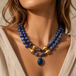 May include: A layered necklace featuring blue beaded strands with gold-colored accents. The necklace includes a pendant with a blue oval stone. The jewelry is displayed on a person wearing a cream-colored top.