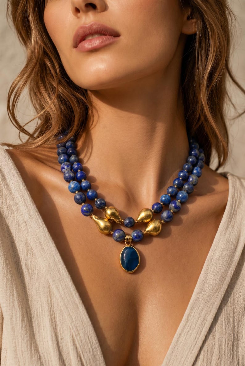 May include: A layered necklace featuring blue beaded strands with gold-colored accents. The necklace includes a pendant with a blue oval stone. The jewelry is displayed on a person wearing a cream-colored top.