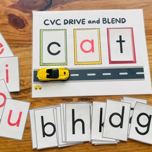 Puede incluir: Un juego educativo CVC Drive and Blend con un coche de juguete amarillo en un camino gris. El juego incluye fichas de letras con las letras e, i, o, u, c, a, t, b, h, l, d y g.