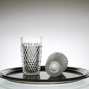 May include: Two clear, diamond-patterned drinking glasses, one upright and one on its side, sit on a silver-colored tray. The glasses have a textured design and are set against a gradient gray background. The tray has a rounded edge.