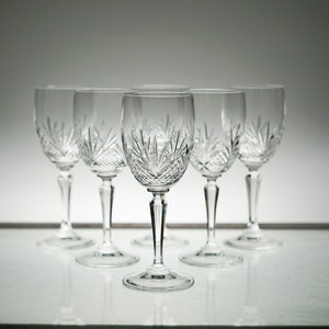 May include: Five clear crystal wine glasses with a cut glass design. The glasses are arranged in a row on a white surface.