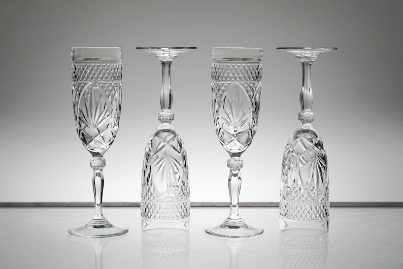 K&ouml;nnte beinhalten: Vier klare Champagnergl&auml;ser aus Glas mit geschliffenem Design. Die Gl&auml;ser sind in einer Reihe auf einer wei&szlig;en Oberfl&auml;che angeordnet.