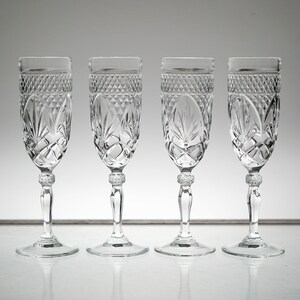K&ouml;nnte beinhalten: Vier klare Champagnergl&auml;ser aus Glas mit geschliffenem Kristallmuster. Die Gl&auml;ser sind in einer Reihe auf einer wei&szlig;en Oberfl&auml;che angeordnet.