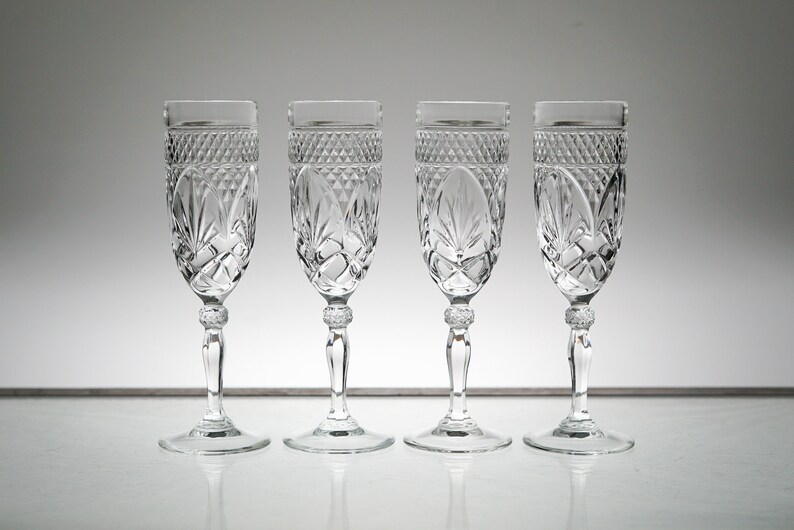 K&ouml;nnte beinhalten: Vier klare Champagnergl&auml;ser aus Glas mit geschliffenem Kristallmuster. Die Gl&auml;ser sind in einer Reihe auf einer wei&szlig;en Oberfl&auml;che angeordnet.
