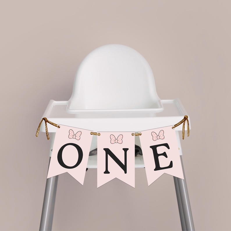 K&ouml;nnte beinhalten: Ein rosa Banner mit dem Wort "ONE" in schwarzen Buchstaben, verziert mit Schleifen, h&auml;ngt an einem Hochstuhl. Der Hochstuhl ist wei&szlig; mit silbernen Beinen, vor einem neutralen Hintergrund. Das Banner ist wahrscheinlich f&uuml;r eine erste Geburtstagsfeier.