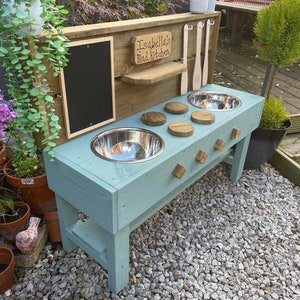 May include: A light blue wooden play kitchen with two stainless steel bowls, wooden utensils, and a chalkboard. The sign reads "Isabella's Mad Kitchen". The play kitchen is outdoors, surrounded by plants and gravel.