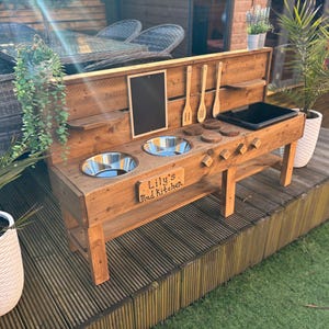 May include: A wooden play kitchen for outdoor use, featuring two stainless steel bowls, a black surface, and wooden utensils. The sign says "Lily's Mad Kitchen". The kitchen is on a wooden deck with potted plants.