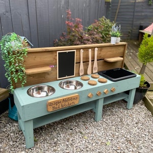 May include: A light blue wooden mud kitchen with two stainless steel bowls, a black tray, and wooden utensils. The sign reads "Unique Abilities Mud kitchen". The outdoor play kitchen is set against a wooden fence, with a gravel surface.