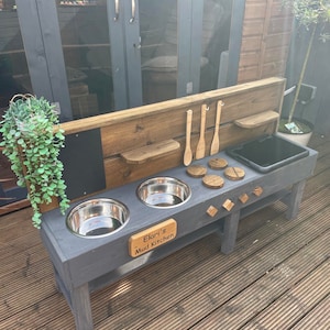 May include: A grey wooden mud kitchen with two stainless steel bowls, a black basin, and wooden utensils. The kitchen has a wooden backsplash with shelves and a small chalkboard. The sign reads "Elari's Mud Kitchen."