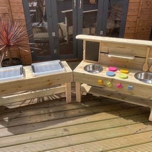 May include: A wooden outdoor play kitchen and a separate wooden table with two clear plastic bins. The play kitchen features two stainless steel sinks, colourful knobs, and wooden utensils. The set is made of light-coloured wood.