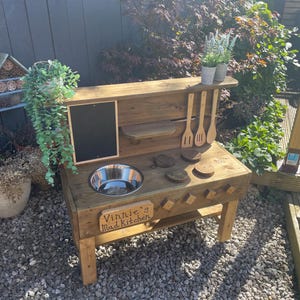 May include: A rustic wooden mud kitchen for outdoor play, featuring a chalkboard, a metal sink, and wooden utensils. The sign reads "Vinnie's Mud Kitchen". The kitchen is surrounded by plants and set on a gravel surface.