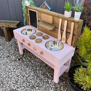 May include: A pink wooden mud kitchen with two stainless steel bowls, wooden utensils, and a chalkboard. The sign reads "Bella's Mud Kitchen". The kitchen is set outdoors with a wooden fence and plants in the background.