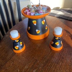 May include: A Halloween-themed candy dish and two candle holders. The black and orange painted ceramic pieces are decorated with candy corn. The candy dish is filled with wrapped lollipops. The candle holders each hold a white candle.