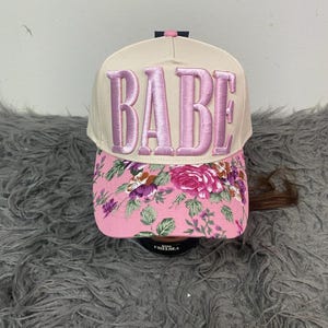 May include: A baseball cap with a cream-colored crown and a pink floral patterned brim. The word "BABE" is embroidered in pink on the front. The cap has a curved brim and a pink bow detail.
