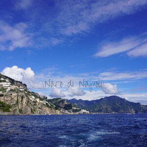 Puede incluir: Un paisaje costero con un mar azul profundo, montañas verdes y un cielo azul brillante con nubes. Se aprecia el texto "Naro di Napoli". La imagen captura una vista pintoresca de la costa.