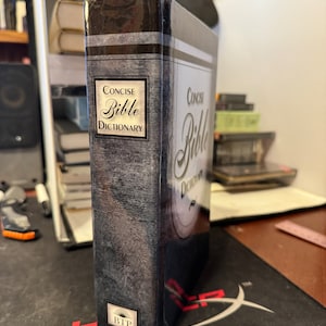 Peut inclure: Un livre noir et gris avec le titre "Concise Bible Dictionary" sur le dos. Le livre a une couverture texturée et un logo argenté en bas.