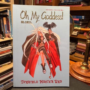 May include: A light blue manga book titled Oh My Goddess! Terrible Master Urd by Kosuke Fujishima. The cover features a long-haired character in a black and red outfit with a black bird perched on a staff, set against a pale blue background.