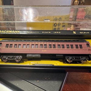 Può includere: Un vagone passeggeri del treno modello Bachmann in scala N in una vetrina. Il vagone è bordeaux con dettagli neri e ruote argentate. La vetrina ha una cornice nera con il logo Bachmann e "Quality Since 1833" stampato sopra.