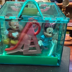 May include: A clear, turquoise plastic carrying case containing Mickey Mouse and Minnie Mouse figurines, along with a red and pink slide. Mickey is wearing brown shorts and Minnie is in a blue dress.