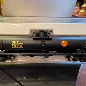 Könnte beinhalten: Ein schwarzer Modell-Eisenbahn-Tankwagen mit Shell-Logo und Text. Der Tankwagen hat eine Leiter und andere Details. Der Text "SHELL SCCX 709" ist sichtbar. Der Modell-Eisenbahnwagen steht auf einem Gleis.