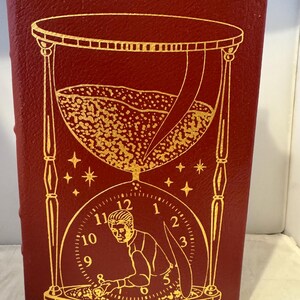 Può includere: Un libro rilegato in pelle rossa con un design a clessidra e orologio dorato. La clessidra è piena di sabbia dorata e il quadrante dell'orologio mostra i numeri da 1 a 12. Una figura è raffigurata all'interno del quadrante dell'orologio, che sembra girare le lancette dell'orologio.