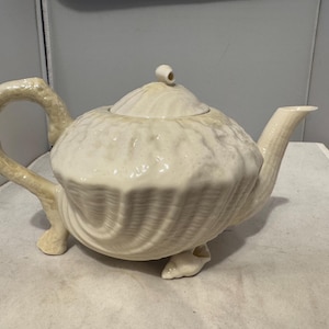 May include: Cream-colored ceramic teapot with a seashell design. The teapot features a rounded body, a curved handle, and a spout. The lid has a small knob. The teapot is resting on three small feet.
