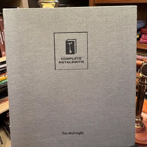 May include: A grey hardcover book titled "COMPLETE METALSMITH" with a square logo featuring a hammer. The author's name, Tim McCreight, is printed below the title. The book is displayed on a dark surface.