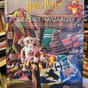 May include: A hardcover book titled Harry Potter Crochet Wizardry, The Official Harry Potter Crochet Pattern Book by Lee Sartori. The cover features a crocheted Dobby figure, a sorting hat, a colorful patchwork blanket, and a phoenix.
