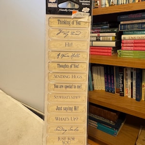 May include: A package of clear epoxy sentiment stickers with phrases like "Thinking of You," "Hi!," and "Sending Hugs." The stickers are rectangular with a cream-colored background and black text. The package also includes the text "Friendly Greetings."