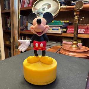 Puede incluir: Figura de Mickey Mouse sonriendo, con chaqueta negra, pantalones cortos rojos y zapatos amarillos. La figura est&aacute; sobre una base amarilla. Una lupa y libros en el fondo.