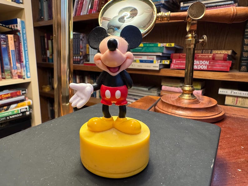 Puede incluir: Figura de Mickey Mouse sonriendo, con chaqueta negra, pantalones cortos rojos y zapatos amarillos. La figura est&aacute; sobre una base amarilla. Una lupa y libros en el fondo.