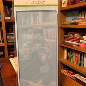 May include: A Cuttlebug cutting pad set in a clear plastic package. The package has a teal and white label with the Cuttlebug logo and text. The cutting pad is a light blue color. The background includes bookshelves and a wooden table.
