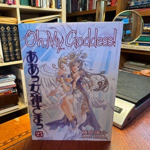 May include: A softcover manga volume titled Oh My Goddess! volume 23 by Kosuke Fujishima. The cover features a light blue and white illustration of two winged figures embracing, with the title text in English and Japanese characters.